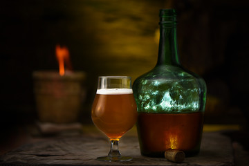 Botella con Cerveza Rubia y copa sobre mesa con fondo iluminado y jarrón con fuego