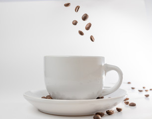 Coffee beans fall into a white porcelain cup and saucer, bouncing and jumping through the air conveying alert excitement, all isolated against a white background
