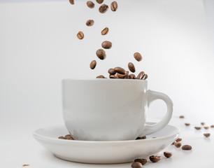 Coffee beans fall into a white porcelain cup and saucer, bouncing and jumping through the air conveying alert excitement, all isolated against a white background