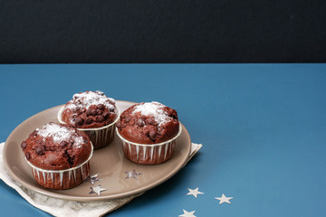 Three chocolate muffins lie on a plate against the background of a turquoise-colored table....