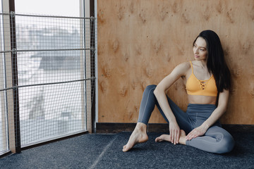 Fototapeta premium Young attractive fitness girl sitting on the floor near the window on the background of a wooden wall, resting on yoga classes