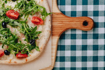 Neapolitan pizza with tuna, cheese, arugula, basil, tomatoes, olives, sprinkled with cheese on a wooden table on a tablecloth in a cell with a place for the text.