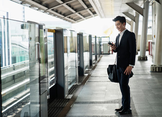 Fototapeta premium Asian executive using smartphone while waiting for the train,Businessman wearing formal blue suit and carry a black bag on shoulder.
