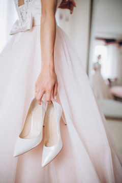 The bride in her wedding dress holds her stylish beige shoes in her hands.