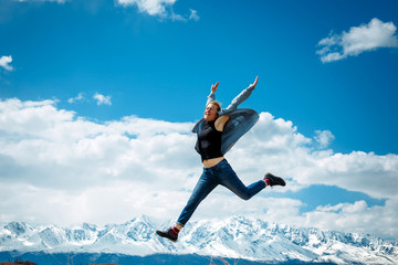 Young woman in jeans jumps high into the air on the background of mountain peaks covered with snow and blue sky with white clouds. Spectacular views of mountain range. Travel and discovery concept.