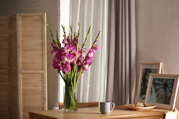 Vase with beautiful pink gladiolus flowers, pictures and cup on wooden table in room, space for text