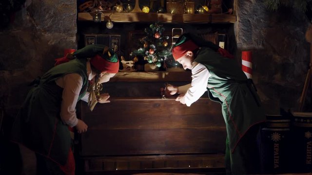 Fairy elves in dark room at Christmas. Excited elves open magic chest and take out a gift indoors. Christmas time.