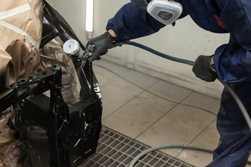 A man in blue overalls and black gloves spray black paint onto a car frame during a body repair in an auto service