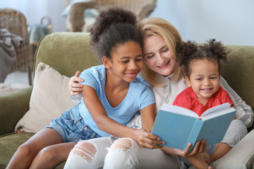 Happy woman with her African-American daughters reading book at home