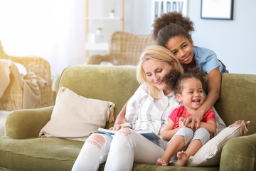 Fototapeta premium Happy woman with her African-American daughters reading book at home