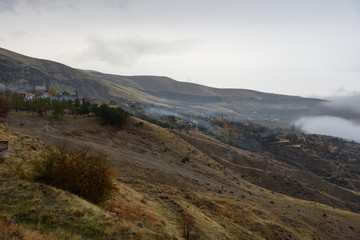 Fototapeta na wymiar Mountains shrouded in mist in the season of golden autumn on a cloudy day, view of the ancient village in the gorge from the top of the mountain.