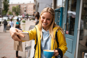 Enchanting blonde young woman with smartphone take selfie on the street