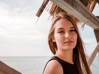 Sexy young woman with beautiful long hair, in an arbor of planks and thatched roof, looks at the sea lit by the sun