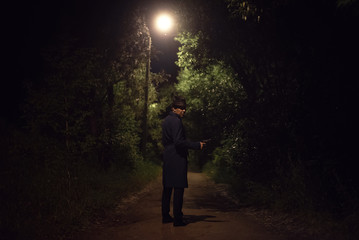 Detective agent in a leather hat and coat is standing and looking into the dark with a handgun in a hand.