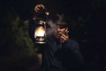 Detective agent in a hat and coat is holding in hand a kerosene lamp and smokes his smoking pipe.