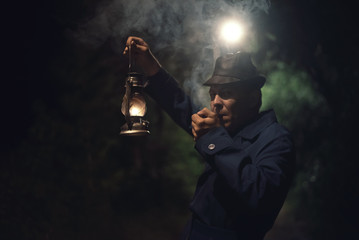 Detective agent in a hat and coat is holding in hand a kerosene lamp and smokes his smoking pipe.