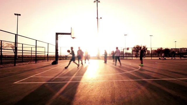 Basketball Match During Sunset shows a group of men playing basketball at sunset. The ground and the sky are enveloped in metallic colors. The men enjoy the games as the continues to set. 