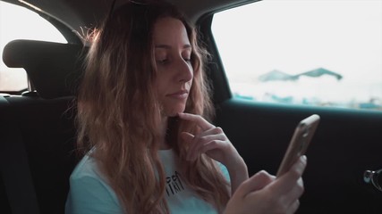 Young woman looking her phone on taxi. On rear seat, beautiful girl interested in mobile phone on road, trip. 