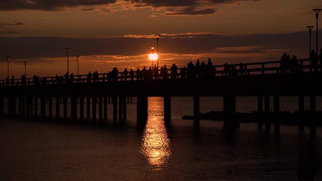 People silhouettes walking along the pier at dramatic sunset on hot summer day in Palanga beach, Lithuania. Colourful sun setting into Baltic sea while tourists enjoying warm weather in popular resort