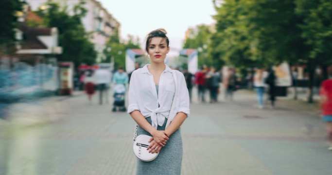 Time lapse of beautiful girl standing outdoors alone looking at camera when men ans women are passing by busy with everyday life. People, lifestyle and youth concept.