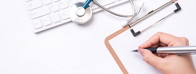 Female doctor writing a medical record case over clipboard on white working table with stethoscope, computer keyboard. Top view, flat lay, copy space