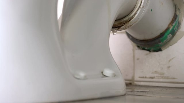 Leaking water from a toilet due to pipe blockage or worn out parts. Smelly water drops dropping behind the toilet from dirty pipe. Toilet leakage, bad smell, plumber needs to take care of it