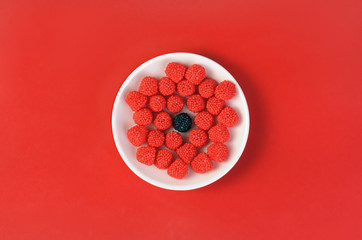 Group of red chewing candies in form of raspberry and one black candy flavored blackberry in the center on a white plate on a red background. Top view.