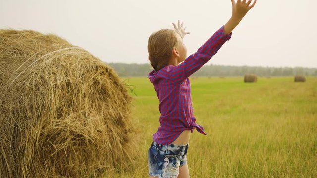 Happy girl stretching hand to sky on hay stack background. Teenager girl raising hands enjoying nature on harvesting field in countryside