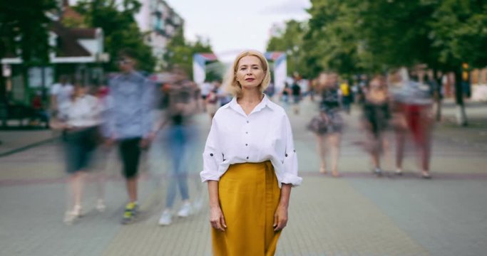 Zoom in time lapse of attractive mature blonde standing in city street on summer day looking at camera alone. Modern lifestyle, people and society concept.