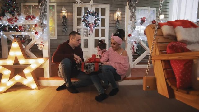 Happy family with little daughter and dog sitting on porch with Christmas tree and decor. Family concept. Young family in pullovers hugging in Christmas day. Wide angle full length shot
