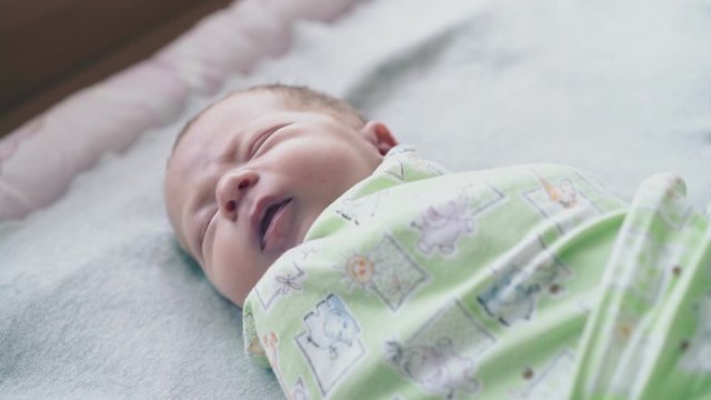 adorable newborn baby lies with closed eyes wrapped in muslin swaddle and starts crying close view slow motion