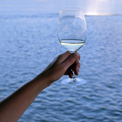 Woman hand holding a glass of white wine against water, sea or lake. Square shape image with copy space.