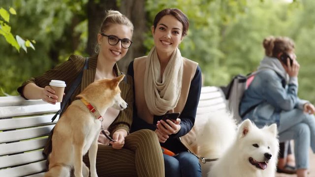 Lockdown of two young multiethnic women sitting with their doggies on bench in park, looking at smartphone screen the looking at camera and smiling