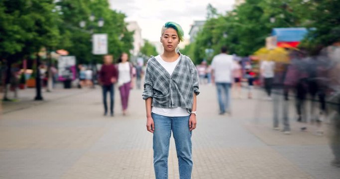 Zoom out time lapse portrait of stylish Asian teen standing in pedestrian street alone and looking at camera with serious face while people are moving around.