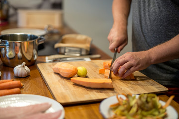 Mann schneidet mit großem Küchenmesser eine Süsskartoffel in Würfel, Knoblauch, Möhren, Fischfilet