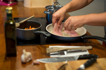 Koch legt mit der Hand Fischfilet in Pfanne am Herd, Töpfe, Ölflasche, Messer, Knoblauch, Nahaufnahme