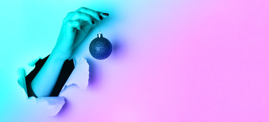 Woman holding red glittering Christmas ball in hand over neon background with snow, light bokeh. Banner, copy space. New year holiday celebration concept