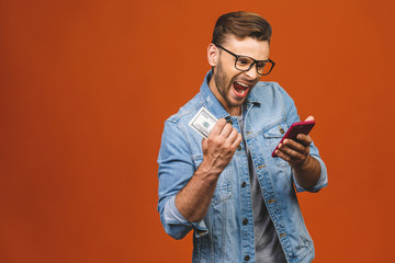 Excited man in casual t-shirt holding lots of money in dollar currencys and using phone in hands isolated over orange wall