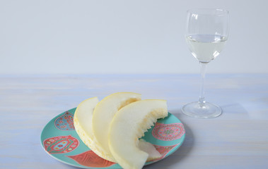 white wine in a glass and ripe yellow sweet melon sliced ​​in a plate with an ornament on a white blue background. Copy space for text.