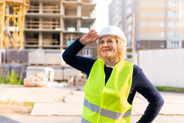 Builder woman at construction site in a green vest and a white helmet with the tablet. Middle-aged woman with glasses looking up.