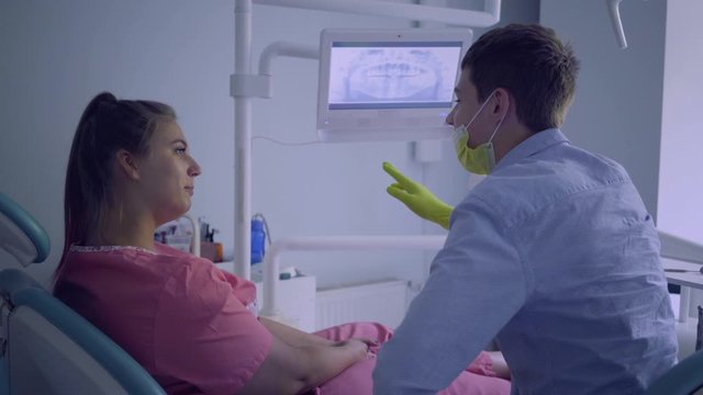 Young man in medical mask showing to his female patient picture of her upper and lower jaws on the screen. The woman visiting the dentist. Dental treatment, medical concept. Dental care.