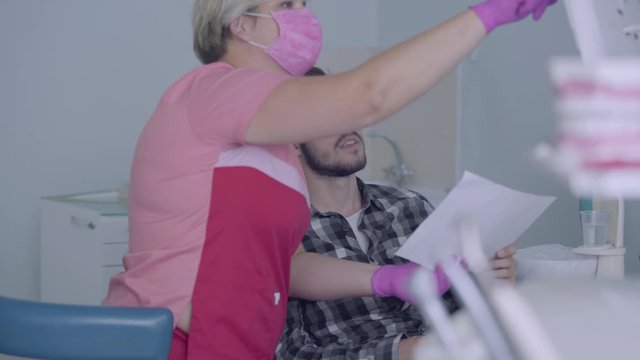 Female dentist in pink mask and gloves showing to male patient picture of his teeth on the screen. The young man visiting the doctor. Dental treatment, medical concept. Dental care.