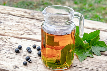 Tea in a glass teapot with green leaves of black currant on a wooden table against the background of green grass.