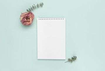 Notepad, sprigs of eucalyptus, dry rose on a pastel turquoise background. Minimalist office desk workspace. Flat lay, top view blank paper copy space mock up.