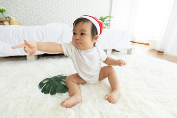 Fototapeta premium Cute Sweet Adorable Asian Baby wearing Santa hat costume sitting on white carpet smiling and playing with happiness emotional in cozy bedroom,Healthy Baby Concept