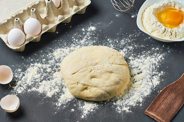 Dough with flour near broken egg with yolk in bowl and other utensil, ingredients lies on dark concrete table. Baking process