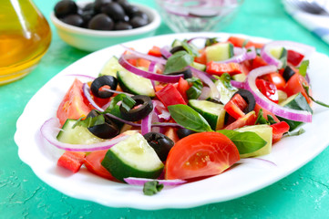 Tasty vitamin vegan salad of tomato, cucumber, red onion, black olives, basil sauce on a white plate on a bright background.
