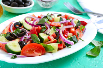 Tasty vitamin vegan salad of tomato, cucumber, red onion, black olives, basil sauce on a white plate on a bright background.