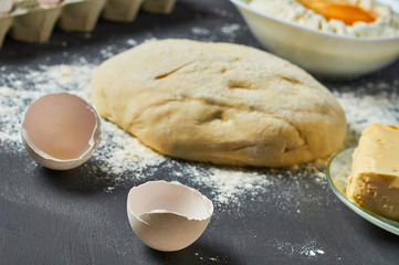 Dough with flour near broken egg with yolk in bowl and other utensil, ingredients lies on dark concrete table. Baking process