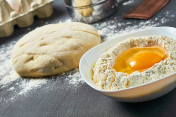 Dough with flour near broken egg with yolk in bowl and other utensil, ingredients lies on dark concrete table. Baking process. Copy space